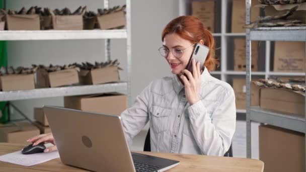 Felice donna piccola imprenditore parlando al telefono e prendendo ordini online in internet store seduto a tavola con computer portatile in magazzino negozio di pesce, sorridente e guardando la fotocamera — Video Stock