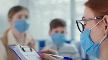 Maskeli ve gözlüklü aile doktoru hastanedeki hasta muayenesi sırasında panoya yazı yazıyor. Annesi ve küçük oğlu doktor randevusuna odaklanamamış.