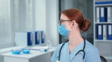 Boynunda steteskop olan mavi üniformalı güzel kadın doktor portresi. Koruyucu maskeli terapist hastanede oturan kameraya bakıyor.