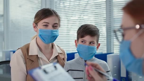 Junge Ärztin macht Notizen und hört Mutter kleiner Patientin zu, maskierte Mutter mit Kind besucht Kinderarzt in Klinik — Stockvideo