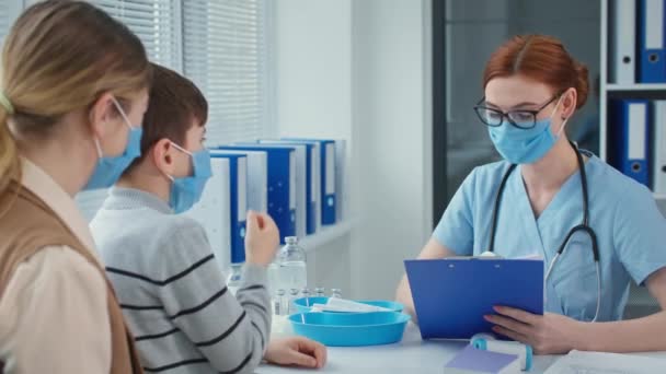 Bra läkare pratar med liten patient sitter i mamma armar, familj läkare i mask och glasögon skriver på Urklipp — Stockvideo