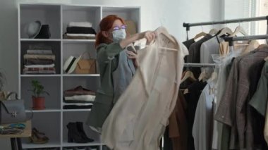 Perakende, tıbbi maske takan genç bayan müşteri mağaza açılışından sonra seviniyor ve alışveriş merkezindeki butiğin açılışından sonra askılardan eşyalar alıyor.