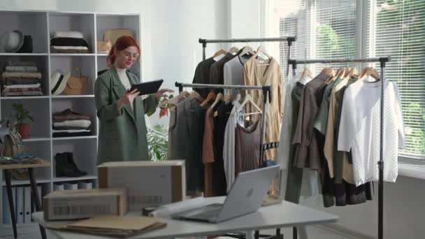 Butik, en ung kvinnlig entreprenör som håller en tablett i handen tar en inventering av kläder på hyllorna i en butik — Stockvideo