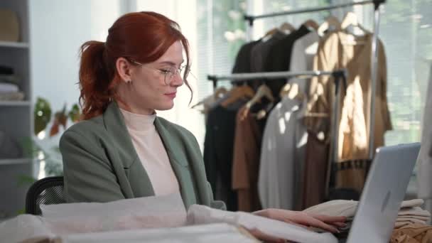 Showroom, bonito vendedor do sexo feminino embalagem de roupas para compradores de lojas on-line sentado em boutique com cabides de roupas no fundo — Vídeo de Stock