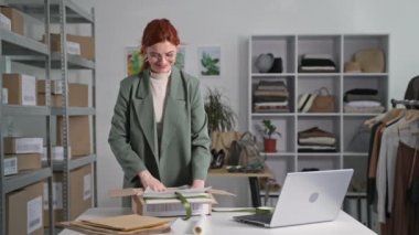 Çevrimiçi perakende satış mağazası, genç iş kadını kıyafetleri bir paket paketliyor bir internet dükkanındaki müşterilere göndermek için dizüstü bilgisayar kullanıyor, gülümsüyor ve kameraya bakıyor.