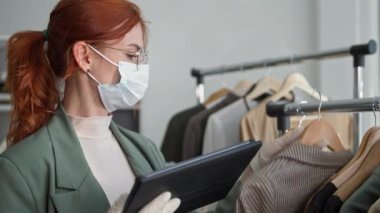 Karantinada küçük bir işletme, tıbbi maskeli ve eldivenli genç bir kadının portresi giyim mağazasında çalışıyor ve tabletteki ürünlerin internetten sipariş verilebilmesi için kontrol ediliyor.