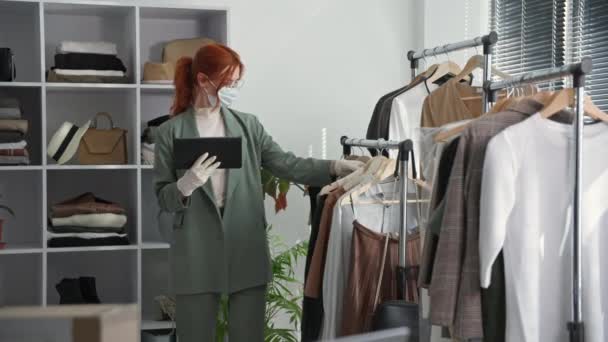 Giyim mağazası, tıbbi maske takan genç bir kadın satıcı ve eldivenler karantinada çalışırken bir online mağazada alıcıların ürünlerini kontrol ediyor. — Stok video