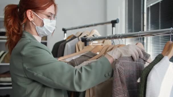 Ung vacker kvinna i en medicinsk mask och handskar iakttar försiktighetsåtgärder tittar på kläder på hängare i en klädaffär efter karantän har lyfts — Stockvideo
