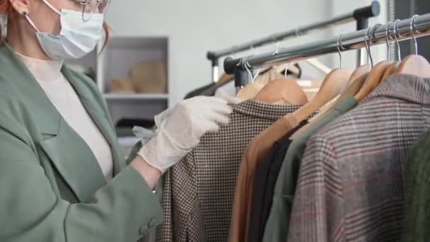 Tienda al por menor, mujer joven en máscara médica y guantes inspecciona perchas con cosas en una tienda de ropa mientras trabaja después de la cuarentena se ha levantado, primer plano — Vídeo de stock