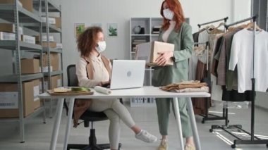 Genç bayan iş kadınları, bir butikte otururken internetteki bir mağazada müşterilere giysi dağıtımı yaparken tıbbi maske takıyorlar.