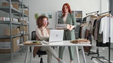 Çevrimiçi perakende, genç kadınlar moda mağazasında depoda otururken müşterileri için internetten giyim mağazalarına bakıyorlar.