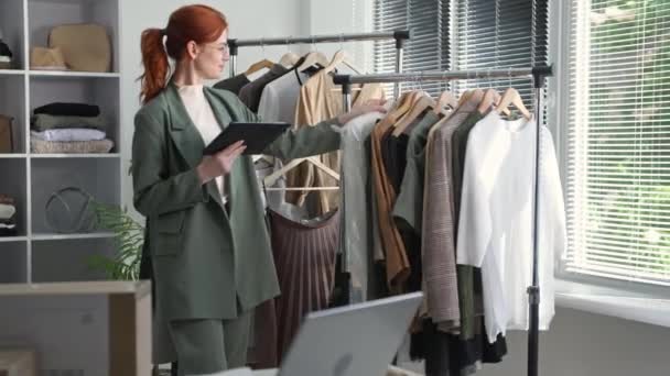 Butik odzieżowy, młoda właścicielka małej firmy w okularach sprawdza dostępność towarów na wieszakach z tabletem w ręku w sklepie odzieżowym — Wideo stockowe