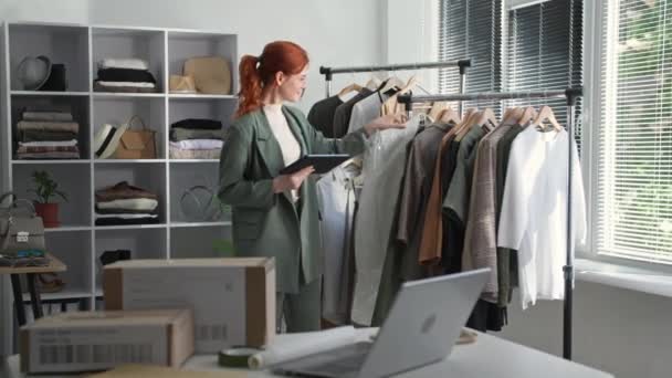 Young business woman owner of clothing store with tablet in hands checks availability of goods on hangers, smiles and looks at camera — Stock Video