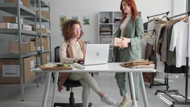 Ung kvinnlig säljare bearbetar internetbeställningar i klädbutik online och gör inköp för kunder tillsammans med kvinnlig kollega sitter i lager i butik — Stockvideo