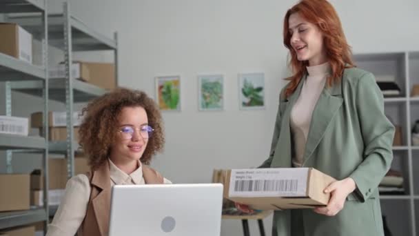 Молода жінка-дизайнер обробляє онлайн-замовлення на ноутбуці для покупців інтернет-магазину одягу, сидячи за столом на складі магазину — стокове відео