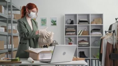 Tıbbi maske ve eldiven takan genç bir iş kadını karantina sırasında müşterilere uzaktan alışveriş ve teslimat ayarlar.