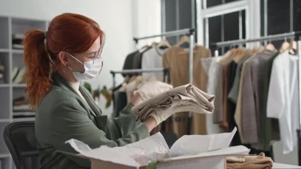 Młoda kobieta w masce medycznej i rękawiczkach organizuje dostawę zakupów w sklepie odzieżowym online i opakowań w pudełku podczas pracy nad kwarantanną — Wideo stockowe
