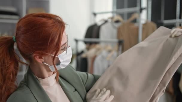 Gözlüklü ve tıbbi maskeli genç bir kadının portresi giysi mağazasında otururken çevrimiçi bir mağazadaki müşterilere teslim edilecek eşyaları kutuya koyuyor. — Stok video