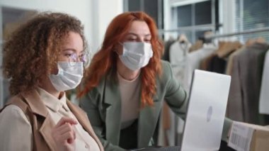 Perakende mağazası, tıbbi maskeli genç bayan yöneticiler elbise askılarının arka planına karşı çevrimiçi bir mağazada müşteriler için bir dizüstü bilgisayar teslimatı ayarlıyorlar.