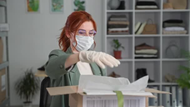 Tıbbi maskeli ve gözlüklü modern genç iş kadını bir depoda otururken bir internet mağazasında müşterilere sergilenmek üzere eşyaları kutuya koyuyor. — Stok video
