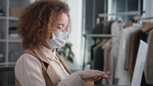 Femme dans le masque médical prépare des marchandises pour les acheteurs dans une boutique en ligne travaillant à l'ordinateur portable avec une collègue femme apportant des boîtes, tout en étant assis dans l'entrepôt du magasin de vêtements — Video