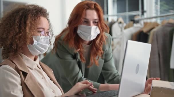 Shopping för köpare, ung säljare i medicinsk mask och glasögon tillsammans med kvinnlig kollega hångla för leverans lådor med bärbar dator när du sitter i nätbutik kläder — Stockvideo