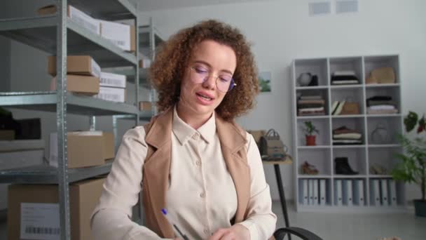 Retrato de una joven gerente que trabaja en la tienda de ropa y se comunica con el cliente a través de video en el portátil, escribiendo orden en blanco mientras está sentada en el almacén de la tienda, sonriendo y mirando a la cámara — Vídeos de Stock