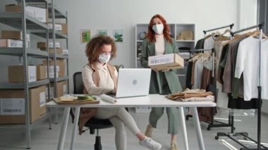 Küçük işletme, tıbbi maskeli çekici kadın işçiler butikte bir depoda duran online bir mağazada mal teslimatı ayarlıyorlar.