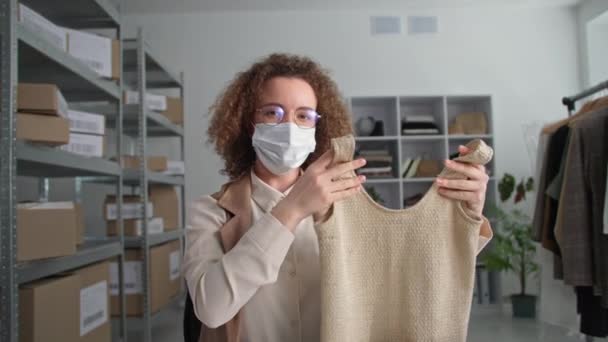 Çevrimiçi ticaret, tıbbi maskeli genç müdürün portresi giyim mağazasında çalışıyor ve bilgisayardaki video bağlantısıyla müşteriyle iletişim kuruyor, mağazadaki malları gösteriyor. — Stok video