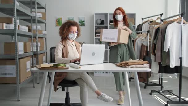 Karantäninköp, unga kvinnliga följeslagare i medicinska masker arbetar i klädbutiken online och ordnar försändelser med varor för leverans till kunder som sitter i lagerbutik — Stockvideo