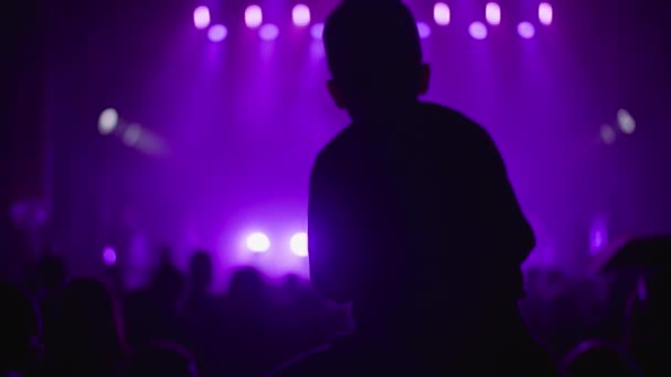 Silhouette fan kid boy on father shoulders enjoying listening to artist during concert at night, people on background of stage with lights in purple color — Stock Video