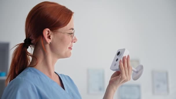 Портрет молодої жінки в окулярах в медичній формі, що тримає приховану камеру спостереження, посміхається і дивиться на камеру — стокове відео