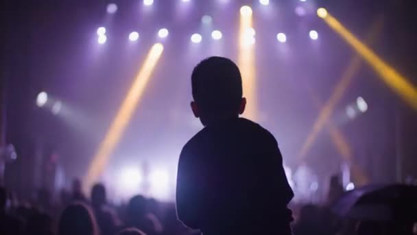 Padre sosteniendo niño en hombros en concierto callejero, multitud de siluetas escuchando la actuación musical en el fondo del escenario con focos — Vídeos de Stock