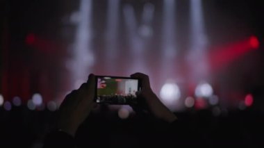Konser, ellerinde cep telefonu olan eller gece çok renkli spot ışıkları olan bir sahnenin videosunu çekiyor.