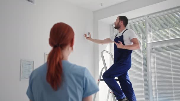 İş güvenliği, genç kadın merdivenlerde erkek uzmanla ofise kamera yerleştirme sürecini izliyor. — Stok video