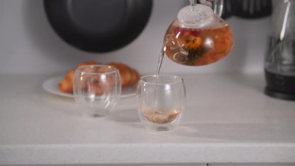 Žena ruka nalévá čaj z průhledné konvice do šálků na pozadí croissanty na talíři, proces vaření čínského čaje květiny — Stock video