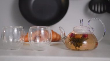 Mutfak masasındaki şeffaf çaydanlıkta çay çiçeği demleme süreci fincanların yanında duruyor, tabakta kruvasanların arka planına karşı, kahvaltı zamanı.
