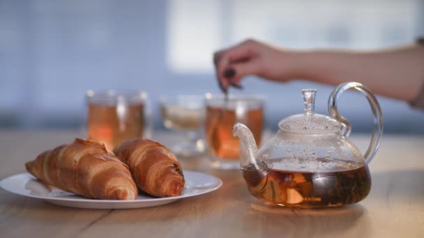 Ketel met een warme drank staat op tafel naast croissants op bord, uit focus vrouwelijke hand giet suiker met een kleine lepel in een kopje met thee — Stockvideo