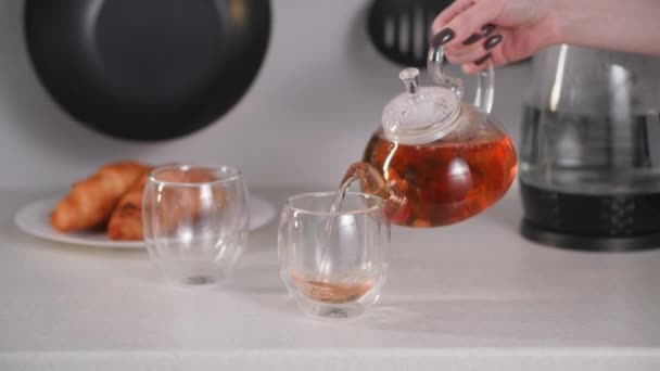 Чайний час, жінка рука наливає гарячий напій з квіткою з прозорого чайника в скляні чашки на тлі круасанів на тарілці — стокове відео