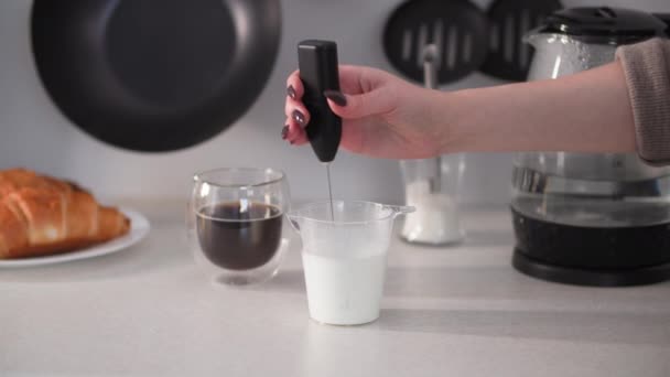 Vaření kávy s mlékem doma v kuchyni, dívka bičuje mléčné výrobky s elektrickou pěnou pak nalévá do průhledného kelímku — Stock video