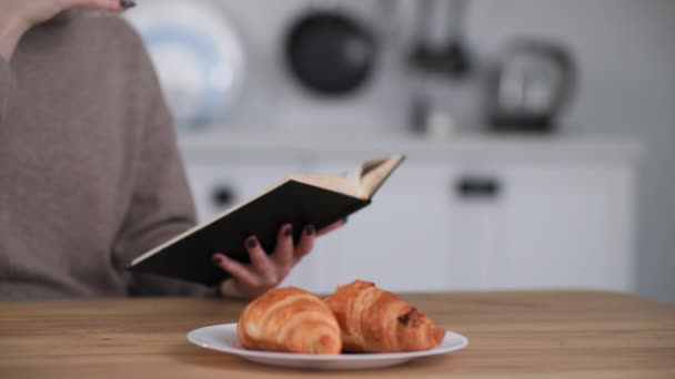 Flicka läsa bok medan du äter frukost sitter vid bordet, oigenkännlig ung kvinna dricka kaffe med mjölk i köket — Stockvideo