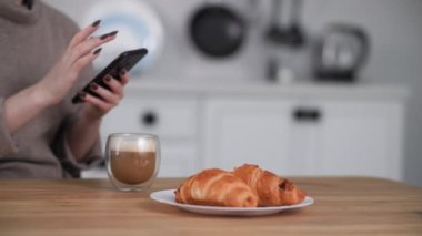 Sabahları mutfakta sütlü kahve içen bir kız, ayçöreği olan bir masada otururken cep telefonu kullanan genç bir kadın.