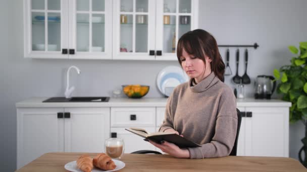 Muße am Morgen, Mädchen liest interessiert ein Buch und trinkt Kaffee mit Milch, während sie zu Hause in der Küche sitzt — Stockvideo
