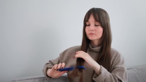 Retrato de menina bonito olhando para a câmera como em um espelho e pentear seu cabelo longo com pente sentado no sofá em casa — Vídeo de Stock