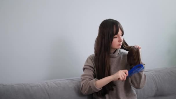 Портрет девушка расчесывает длинные каштановые волосы, сидя на диване перед камерой, молодая женщина делает укладку дома — стоковое видео