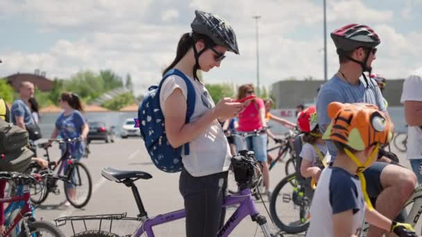 Херсон, Украина 10 августа 2021 года: молодая женщина в шлеме и очках пользуется мобильным телефоном для просмотра маршрута на фоне толпы людей на велосипедах во время езды на велосипеде — стоковое видео