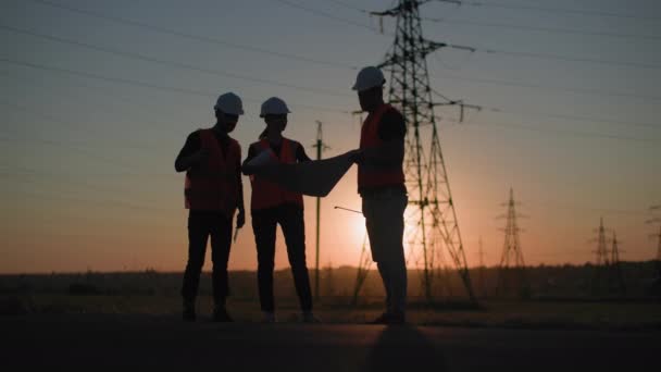 Hombres y mujeres profesionales con el proyecto en sus manos están discutiendo la estructura de las líneas de una torre eléctrica en el fondo de la puesta del sol, silueta — Vídeos de Stock
