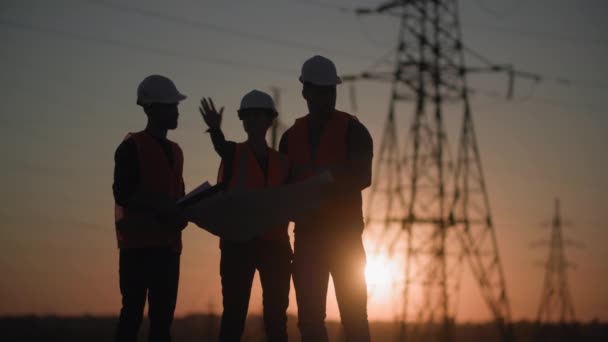 Equipo de especialistas en la construcción de redes eléctricas están discutiendo proyecto de torres de energía fondo de la planta de energía y puesta de sol, silueta — Vídeo de stock