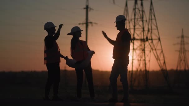Línea eléctrica, mujeres y hombres ingenieros de montaje saltando levantando las manos regocijándose en el exitoso proyecto de construcción de torres de energía para el telón de fondo de la ciudad de puesta de sol, silueta — Vídeo de stock