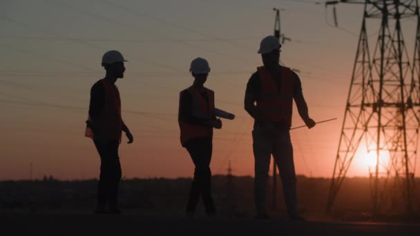 Construção de usina, especialistas da indústria feminina e masculina em capacetes de proteção discutindo a instalação de linha electro para fornecer eletricidade para a cidade ao pôr do sol, silhueta — Vídeo de Stock